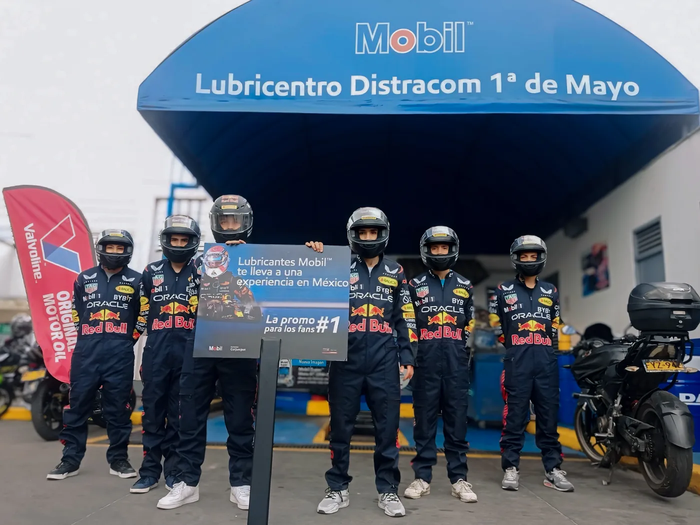 Equipo promocional vestido como pilotos de carreras para activación Mobil en lubricentro, con señalización de campaña y motocicleta