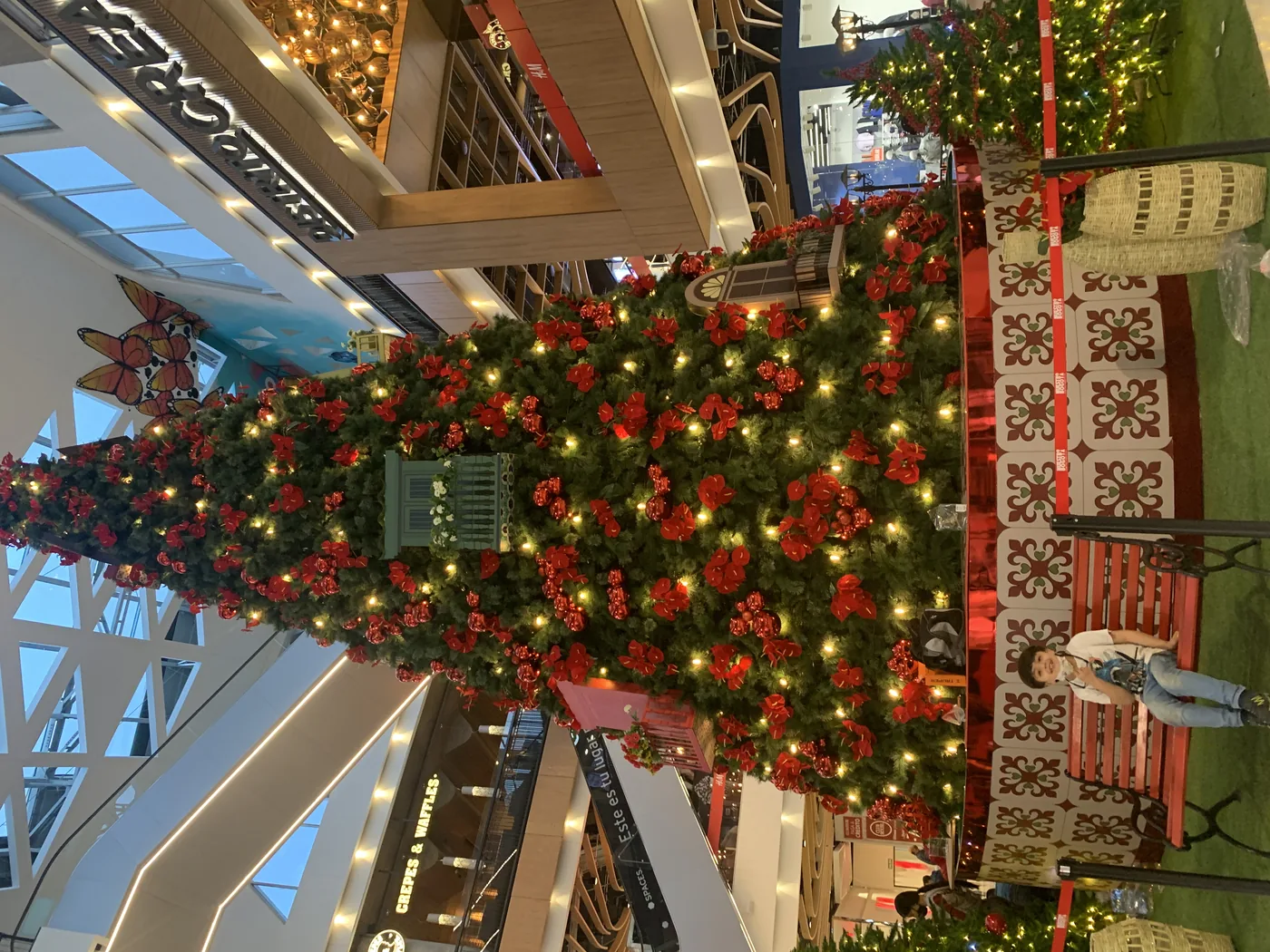 Árbol navideño de gran altura en centro comercial con decoración roja, iluminación cálida, elementos escenográficos y ambientación interior de temporada