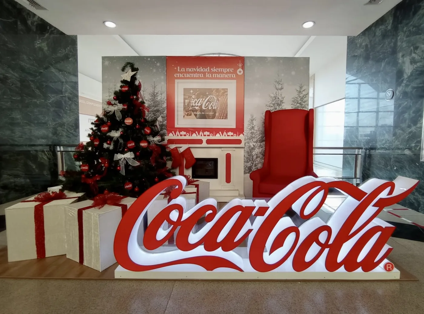 Escenografía navideña de Coca-Cola con árbol decorado, chimenea temática, silla roja, regalos gigantes y logotipo volumétrico iluminado