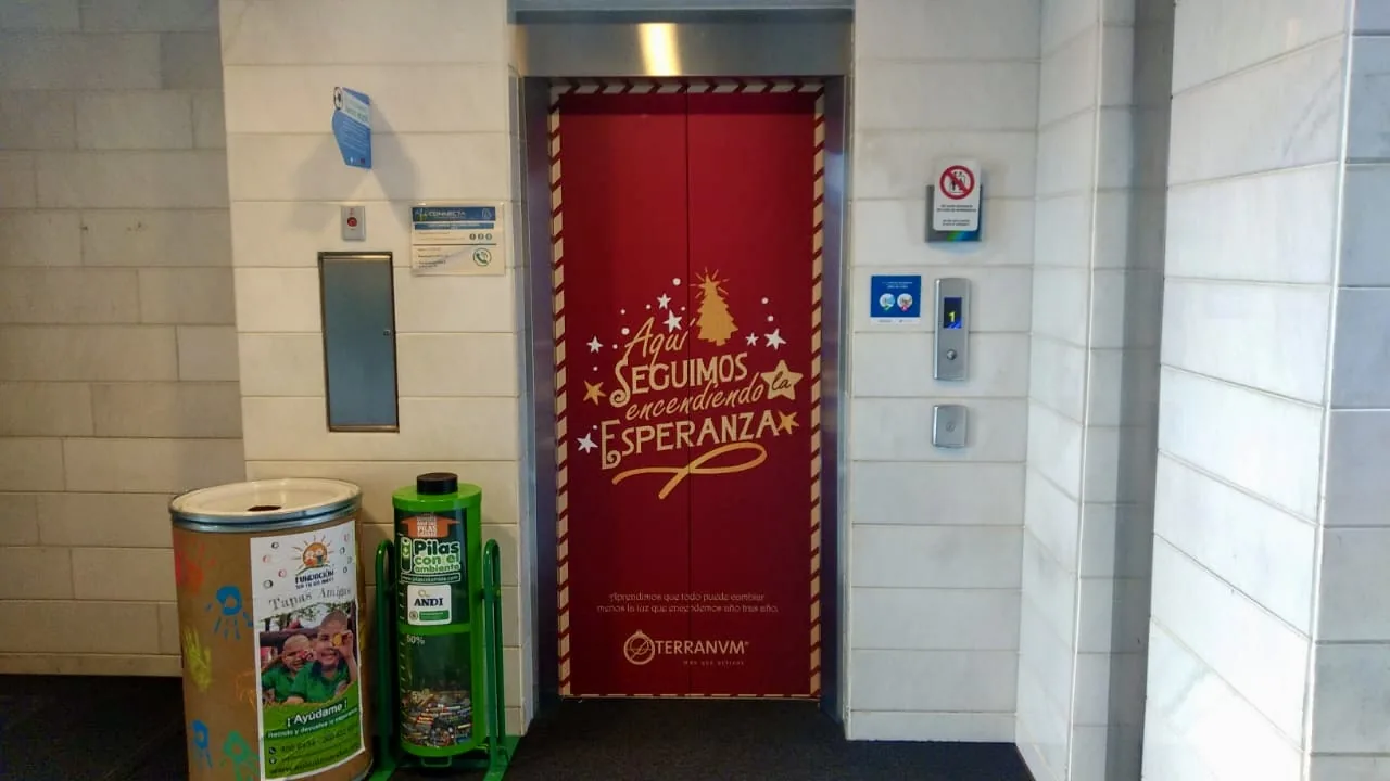 Intervención gráfica navideña en puertas de ascensor con mensaje de temporada, color rojo corporativo y diseño institucional