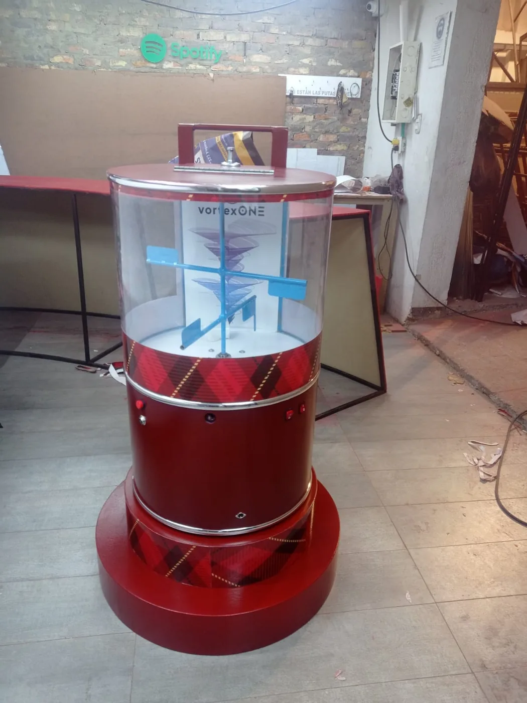 Elemento interactivo cilíndrico tipo vórtice navideño con base roja, cubierta transparente, sistema giratorio interno y acabado decorativo de temporada