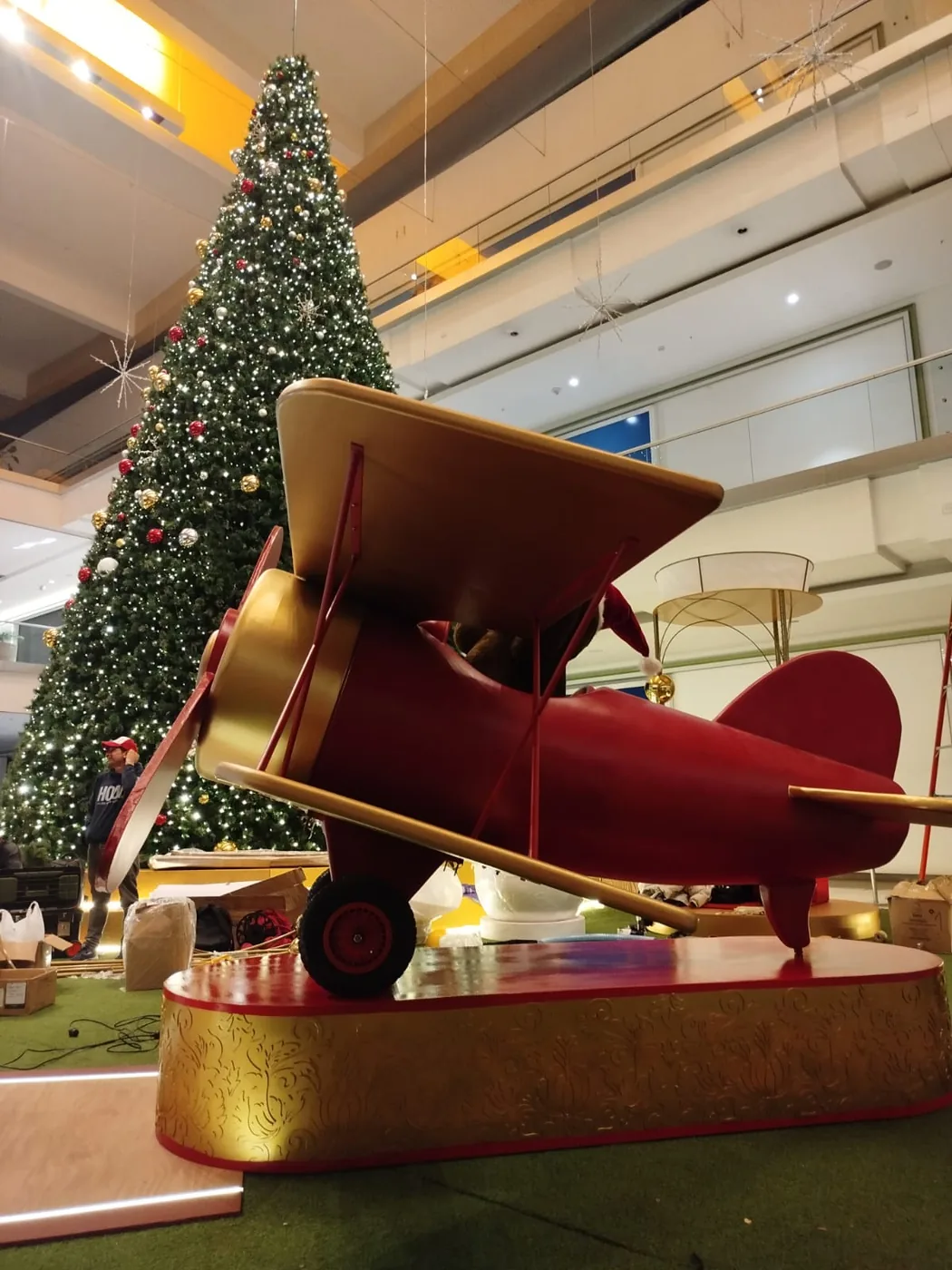 Árbol de Navidad en centro comercial Multiplaza con decoración de gran formato, iluminación cálida y elemento aéreo tipo avioneta como parte de la escenografía