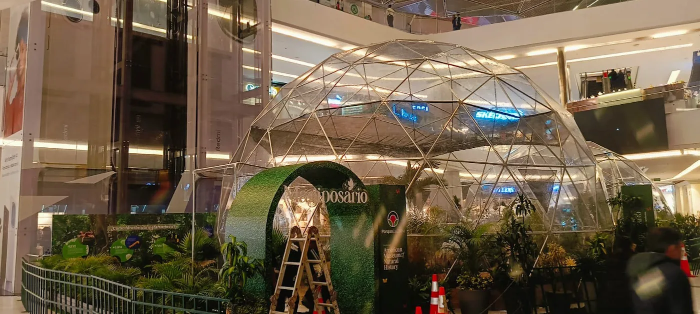 Elemento escenográfico de mariposa monumental instalado en espacio temático de centro comercial, acompañado de ambientación vegetal y estructura geodésica